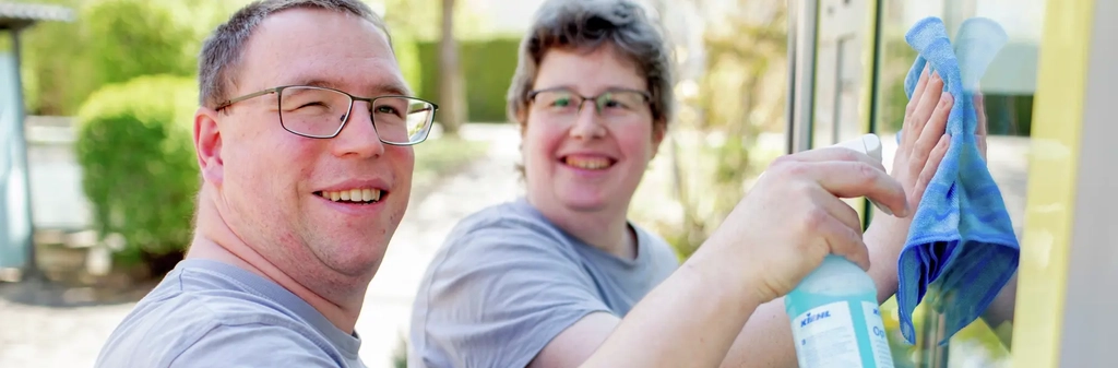 Zwei Personenputzen mit Sprühflasche und Putzlappen eine Fensterscheibe. Beide lachen.