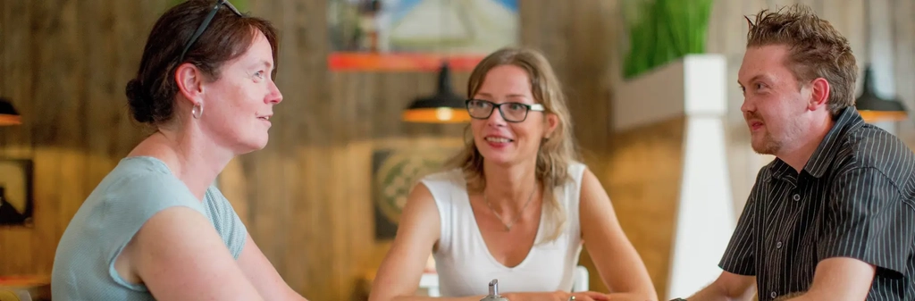 Zwei Frauen und ein Mann sitzen um einen Tisch und lächeln sich an.