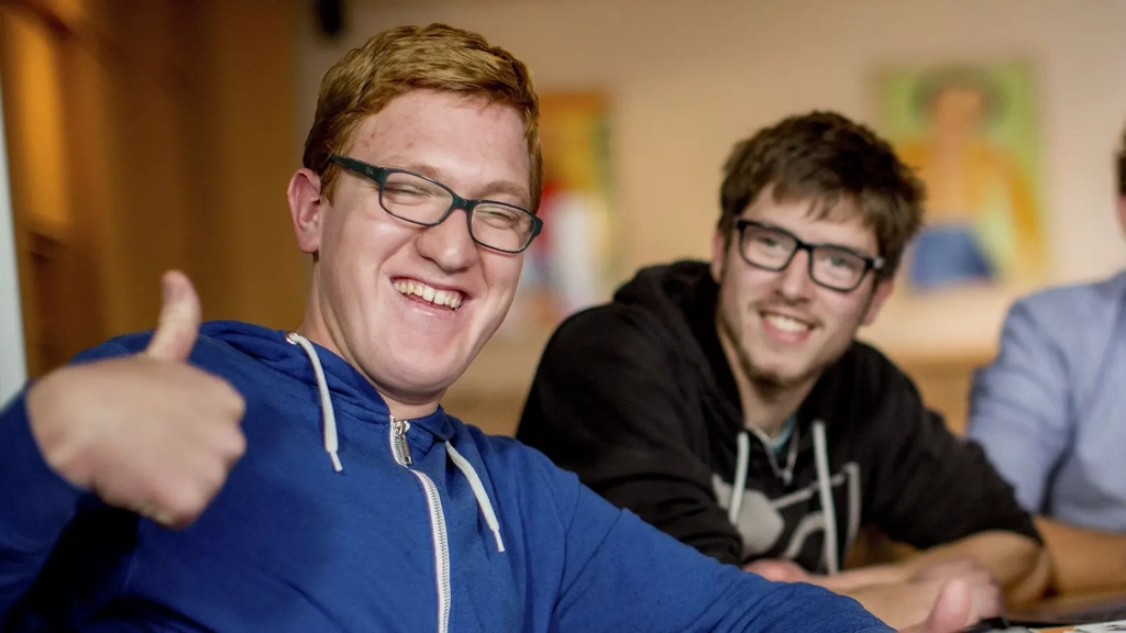 Drei Männer mit und ohne Behinderung sitzen an einem Schreibtisch vor zwei Laptops. Einer von ihnen gibt das Daumen-hoch-Zeichen. Alle drei lachen.