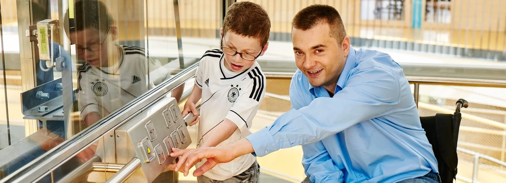 Ein Junge und ein Mann im Rollstuhl drücken die Zahl eines Fahrstuhls.
