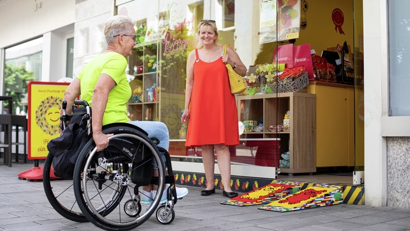 Rita benutzt eine ihrer selbstgebauten Legorampen, um mit ihrem Rollstuhl in einen Laden zu fahren. Die Ladenbesitzerin steht daneben und lächelt.