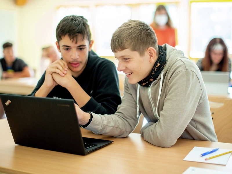 Zwei Jugendliche sitzen nebeneinander in einem Schulungsraum. Sie schauen gemeinsam auf einen Laptop-Bildschirm.