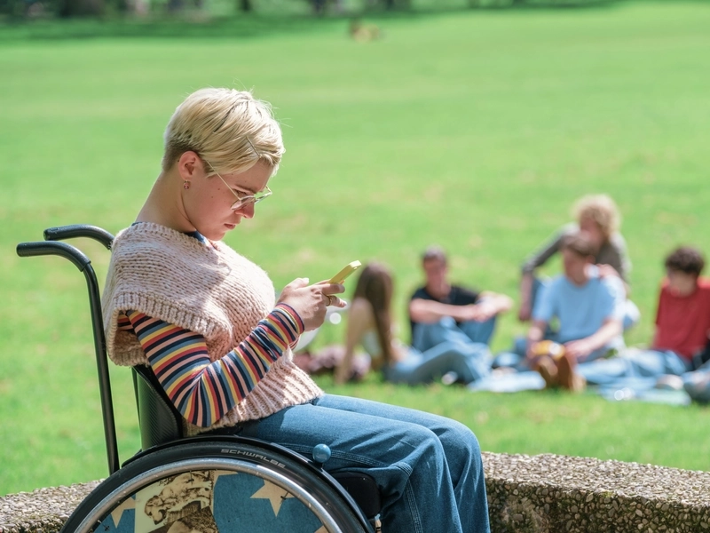 Junge Frau mit kurzen blonden Haaren steht mit ihrem Rollstuhl vor einer kleinen Mauer in einem Park und guckt ins Handy. Auf der Wiese im HIntergrund jenseits der Mauer eine Gruppe anderer junger Leute.