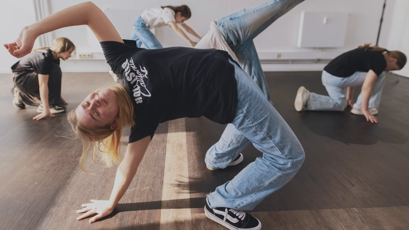 Fünf Mädchen im Teenager-Alter sind in verschiedenen, teils akrobatischen Haltungspositionen in einem leeren Raum verteilt. Es sieht aus, als ob sie eine Tanz-Choreografie ausführen. Alle tragen lange blonde Haare, Jeans, T-Shirts und Sneakers.