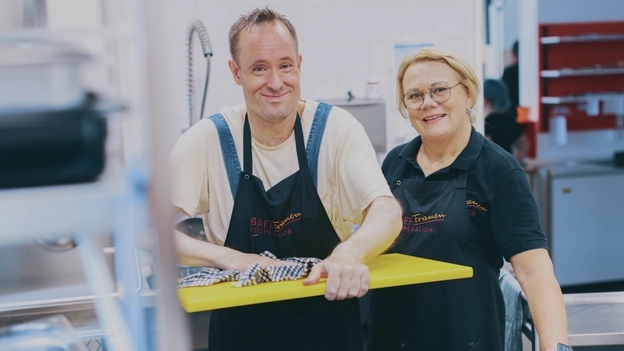 Ein Mann und eine Frau in schwarzer Schürzen stehen in einer Küche. Der Mann trocknet ein gelbes Brett ab.