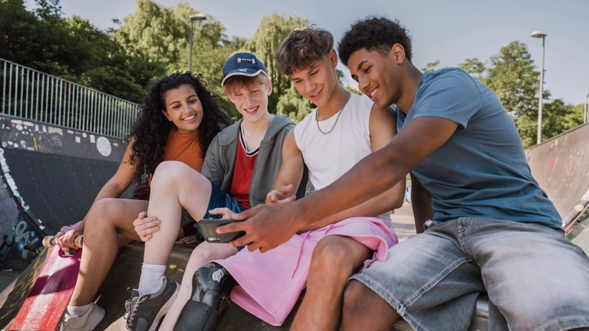 Vier Jugendliche in sommerlicher Kleidung sitzen nebeneinander auf einer Skate-Rampe.  Sie schauen gemeinsam auf ein Smartphone und lachen. Einer der Jugendlichen trägt eine Beinprothese.
