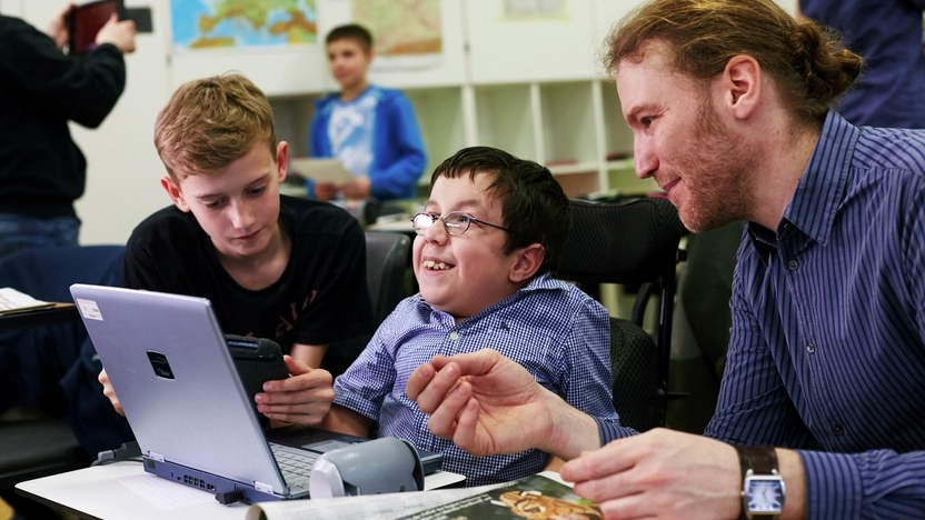 Zwei Schüler mit und ohne Behinderung sitzen gemeinsam mit einer Lehrkraft im Klassenzimmer vor einem Notebook.