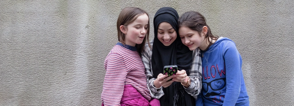 Drei Mädchen stehen nebeneinander vor einer Beton-Wand. Sie alle schauen auf das Handy, das das Mädchen in der Mitte in der Hand hält.