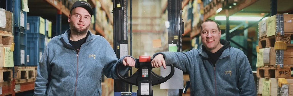 Zwei junge Männer stehen neben einem Gabelstapler in einer Lagerhalle.