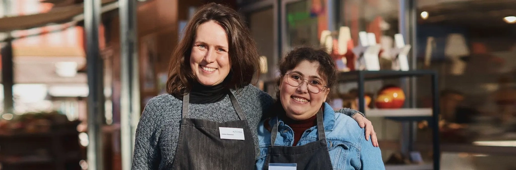 Zwei Frauen mit dunklen Schürzen stehen vor einem Marktstand. Eine hat den Arm freundschaftlich um die Schulter der anderen gelegt und beide lächeln in die Kamera.