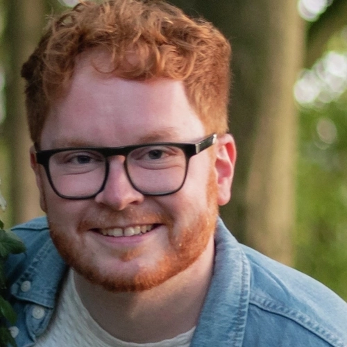 Porträt eines Mannes mit rotem Haar, Dreitagebart und Brille. Er trägt ein Jeanshemd und lächelt in die Kamera.