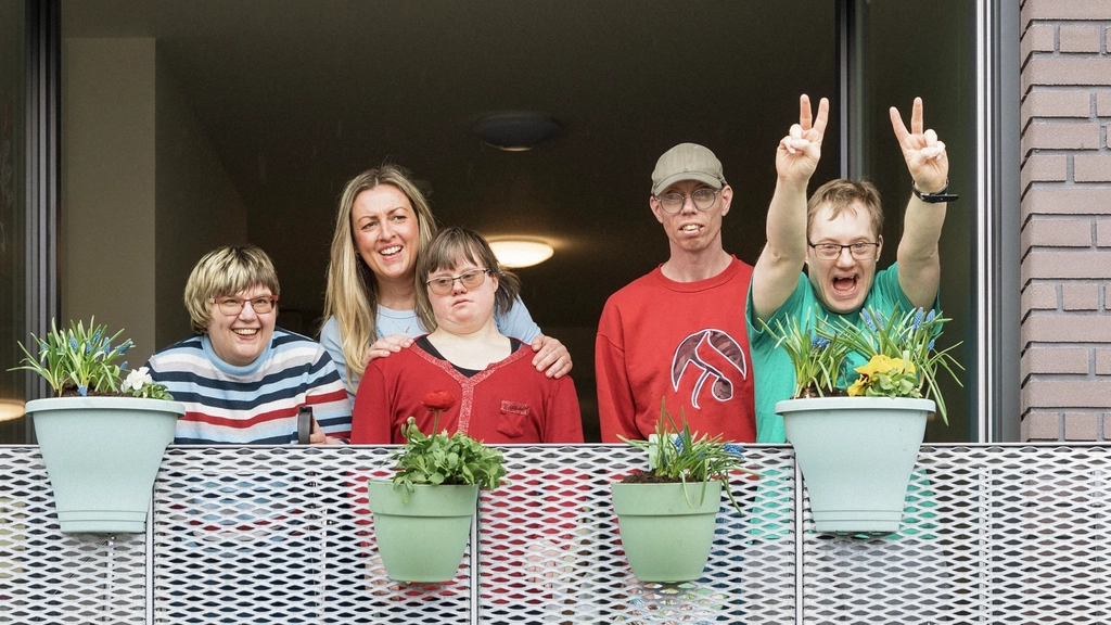 Eine Gruppe von Menschen mit und ohne Behinderung steht auf dem Balkon einer Wohnung. Sie lachen und winken der Kamera zu.