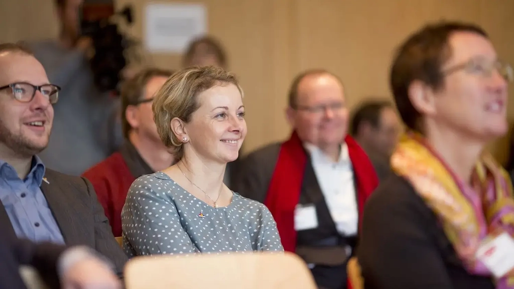 Teilnehmer*innen einer Konferenz sitzen in Stuhlreihen und schauen interessiert nach vorne.