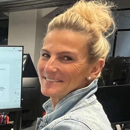 Eine Frau mit blondem Dutt und Jeansjacke steht an einem Schreibtisch. Sie lächelt in die Kamera.