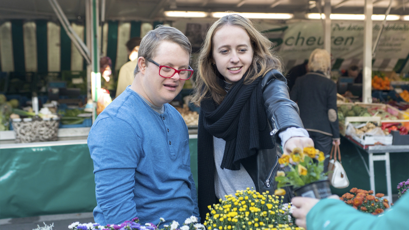 Junger Mann mit Down-Syndrom und eine junge Frau betrachten Blumen vor einem Marktstand