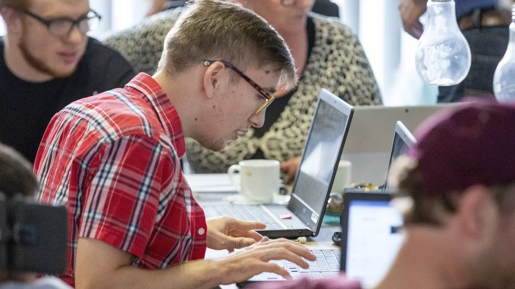 Ein junger Mann arbeitet an einem Laptop. Um ihn herum sitzen weitere Personen mit und ohne Behinderung und arbeiten ebenfalls.