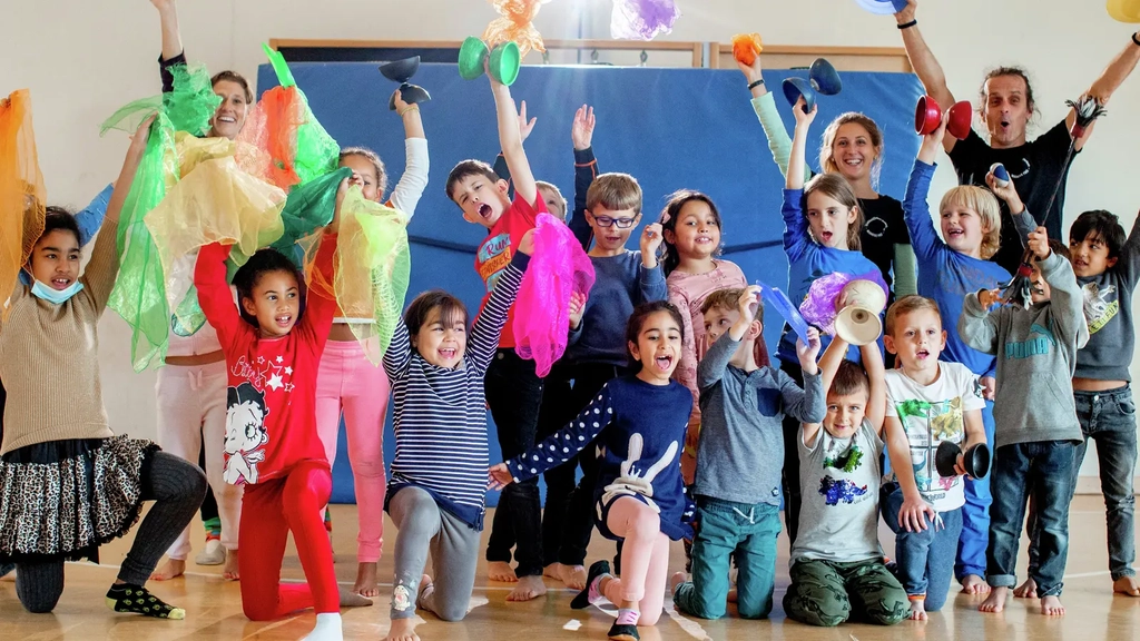 Eine Gruppe von Kindern in einer Turnhalle. Alle jubeln und halten bunte Tücher, Diabolos und Bälle in die Luft.