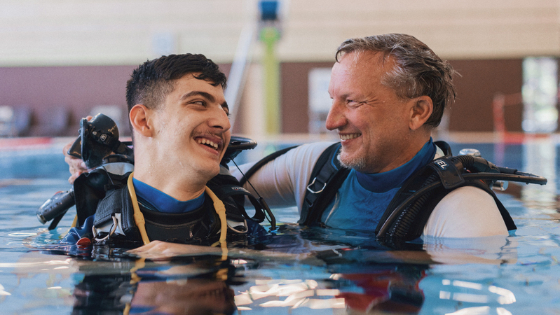 In einem Schwimmbecken befinden sich ein junger und ein älterer Mann. Sie beide tragen Tauchausrüstung. Sie schauen einander an und lächeln sich fröhlich zu.