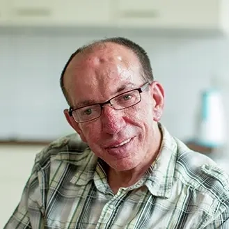 Älterer Mann mit Brille in Jeans und kariretem Hemd sitzt auf dem blau bezogenen Bett seines Appartements und lächelt in die Kamera