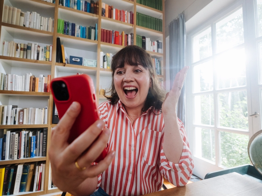 Eine Frau in einem rot-weiß gestreiften Hemd sitzt an einem Tisch in einem hellen Raum und hält ein rotes Smartphone in der Hand. Sie freut sich offensichtlich über etwas und gestikuliert mit ihrer freien Hand, um ihre Emotionen auszudrücken. Neben ihr auf dem Tisch steht ein Globus.