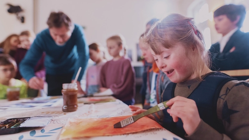 In einem großen hellen Raum sitzen und stehen rund ein Dutzend Kinder um einen Maltisch herum. Eine erwachsene Person beugt sich auch über den Tisch und malt. Das Foto ist von der Höhe der Tischplatte aus aufgenommen. Alle Personen sind nur unscharf zu erkennen, bis auf ein Mädchen gleich im Vordergrund. Sie hat eine sichtbare genetisch bedingte Beeinträchtigung. Mit Daumen und Zeigefinger führt sie einen Pinsel in der linken Hand, mit dem sie Wasserfarbe auf ein Blatt aufträgt. Ihre langen blonden Haare sind mit einer Klammer hinterm Kopf zusammengehalten..
