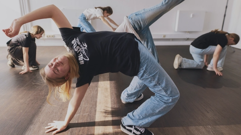 Fünf Mädchen im Teenager-Alter sind in verschiedenen, teils akrobatischen Haltungspositionen in einem leeren Raum verteilt. Es sieht aus, als ob sie eine Tanz-Choreografie ausführen. Alle tragen lange blonde  Haare, Jeans, T-Shirts und Sneakers.