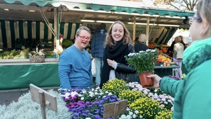Ein junger Mann und eine junge Frau schauen sich Blumen an einem Marktstand an. Der junge Mann hat das Down-Syndrom.