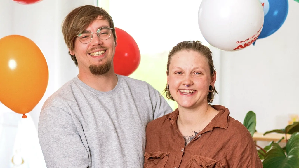 Ein junges Paar steht lachend nebeneinander. Sie haben beide eine Hand auf den schwangeren Bauch der Frau gelegt. Um sie herum fliegen bunte Luftballons.