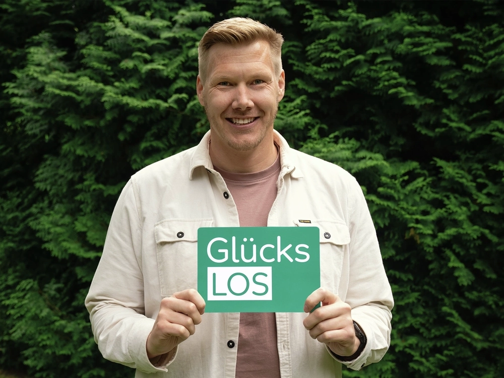 Der Gewinner Daniel Kater steht in einem Garten und lächelt in die Kamera. In den Händen hält er vor dem Körper das grüne Glücks-Los der Aktion Mensch.