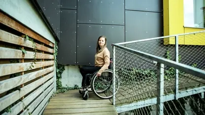 Eine junge Frau im Rollstuhl fährt eine Rampe an einem Haus runter.
