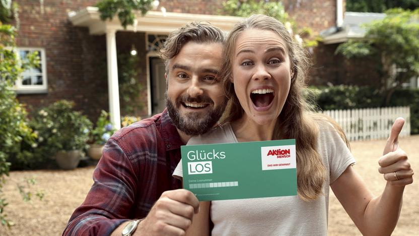 Ein Mann und eine Frau legen die Köpfe aneinander und lachen jubelnd. Sie halten ein Glückslos in die Kamera.