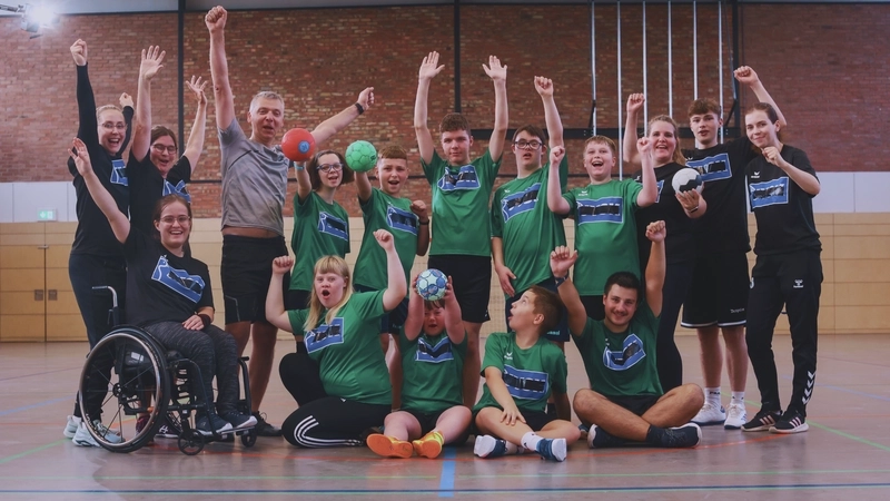 Mannschaftsfoto eines inklusiven Handballtaems, bestehend aus jugendlichen Jungen und Mädchen mit und ohne Behinderung. Si etragen schwarze oder grüne Trikots. Ein Teil von ihnen sitzt auf dem Hallenboden, die anderen stehen dahinter in der zweiten Reihe. Alle recken die Arme.