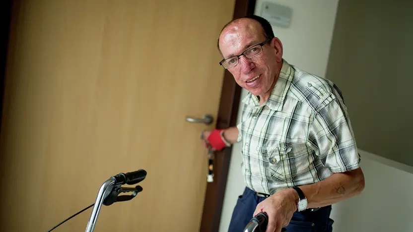 Älterer Mann in Jeans und kariertem Hemd verlässt mit seinem Rollator die Wohnung und schließt hinter sich die Tür.