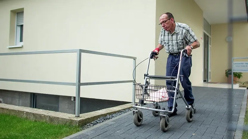 älterer Mann in Jeans und kariertem Hemd verlässt mit Rollator ein Mehrparteienhaus über eine Rampe