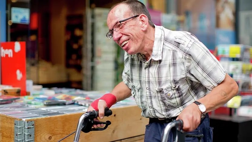 Älterer Mann mit Brille in Jeans und kariertem Hemd geht mit seinem Rollator an einem Geschäft in der Fußgängerzone vorbei und lacht glücklich.