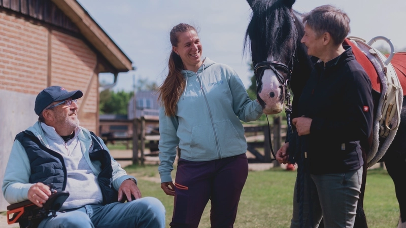 Draußen auf dem Gelände eines Reithofs: Ein älterer Mann im Rollstuhl unterhält sich mit zwei Frauen, die ein schwarzes Pferd in ihrer Mitte am Zaumzeug halten.