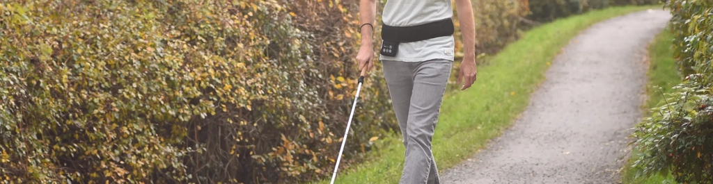 Ein Mann mit Blindenstock geht auf einem schmalen Weg zwischen zwei Hecken. Um seine Hüfte trägt er einen breiten schwarzen Gürtel mit Bedienfeld.