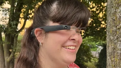 Junge Frau mit langen dunklen Haaren und sommerlicher roter Bluse trägt ein Brillengestell, in dessen rechten Bügel eine Kamera, ein kleines Display und weitere Technik integriert sind.