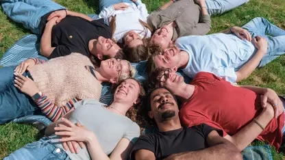 Eine diverse Gruppe von acht älteren Jugendlichen liegt sternförmig im Kreis angeordnet auf dem Rücken im Gras. Alle haben die Augen geschlossen und lachen.