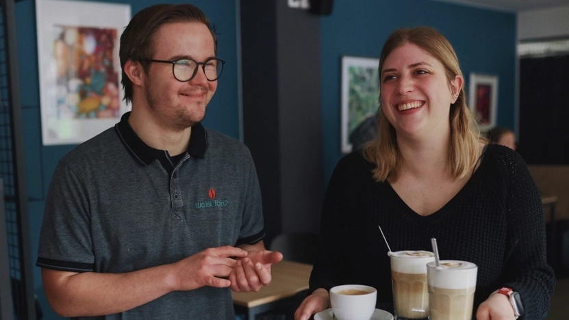 Ein junger Mann mit Downsyndrom und eine junge Frau, die ein Tablett mit Kaffeetassen trägt, stehen lächelnd nebeneinander