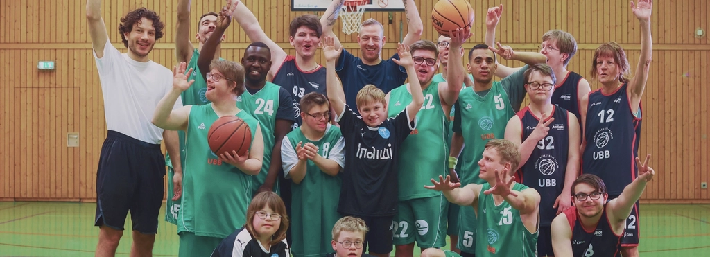 Etwa 20 junge Menschen in grünen und blauen Basketballtrikots haben sich zu einem Gruppenfoto im Mittelkreis einer Sporthalle aufgestellt. Die Jüngsten sind unter 10, die Ältesten etwa 30 Jahre alt. Alle winken in die Kamera.