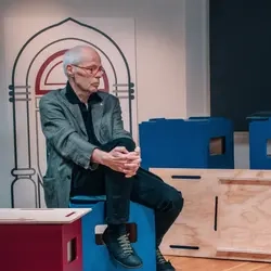 Ein Mann im höheren Erwachsenenalter sitzt in einem Filmstudio auf einem Hocker