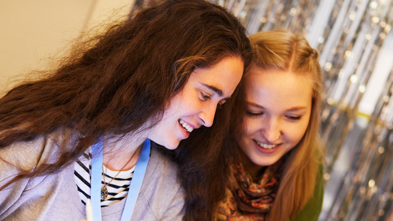 Zwei jugendliche Frauen mit langen Haaren sitzen nebeneinander. Eine hält ein Tablet in der Hand, die andere ein Smartphone auf das sie beide schauen.