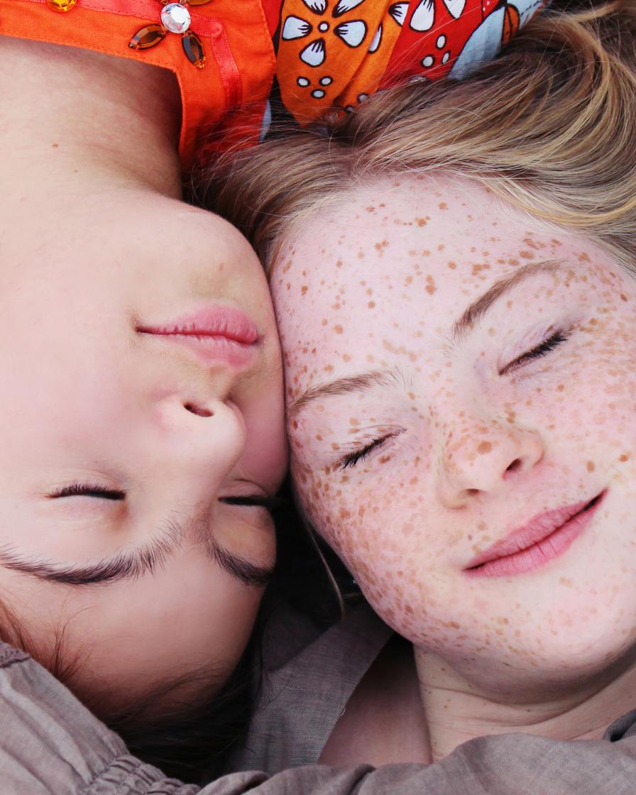 Blick von oben: Zwei Mädchen liegen mit ihren Köpfen dicht beieinander. Sie haben die Augen geschlossen und lächeln zufrieden.