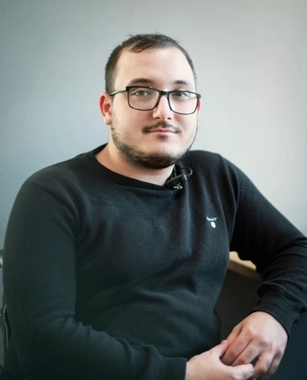 Ein junger Mann mit Brille, dunklen kurzen Haaren und Vollbart in schwarzem Pullt sitzt an einem Tisch und lächelt in die Kamera.