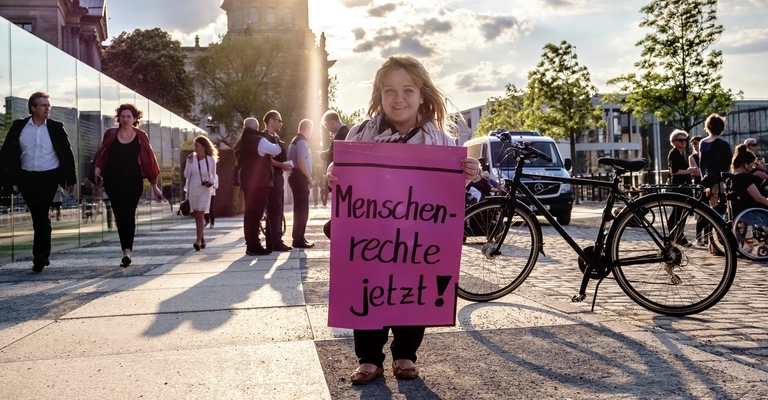 Eine kleinwüchsige Frau hält im Abendlicht ein Plakat mit der Forderung: Menschenrechte jetzt!