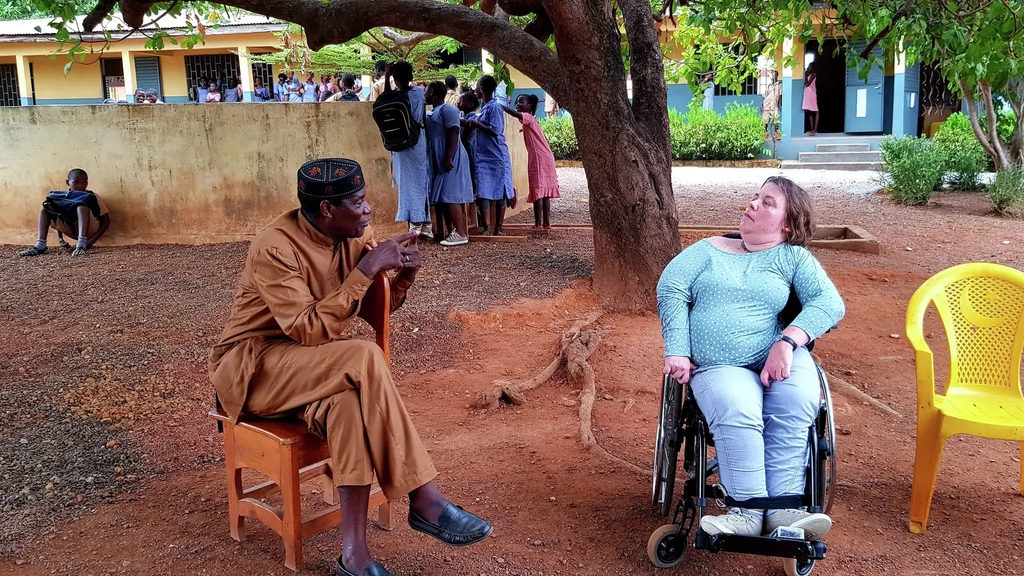Eine Frau im Rollstuhl und ein schwarzer Mann in traditioneller Kleidung im Gespräch unter einem Baum vor einer afrikanischen Schule.