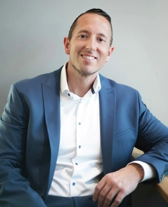 Ein junger Mann mit kurzen dunklen Haaren in blauem Anzug und weißem Hemd steht vor dem Supermarkt, den er managt und strahlt in die Kamera.