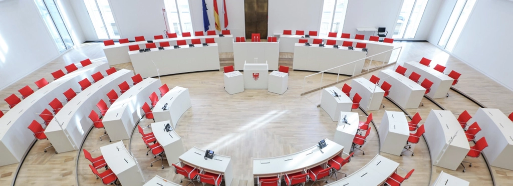 Blick von der Besuchertribühne auf den Plenarsaal. Im Halbkreis angeordnete weiße Abgeordnetenbänke mit roten Stühlen. Am Rednerpult der Wappenadler