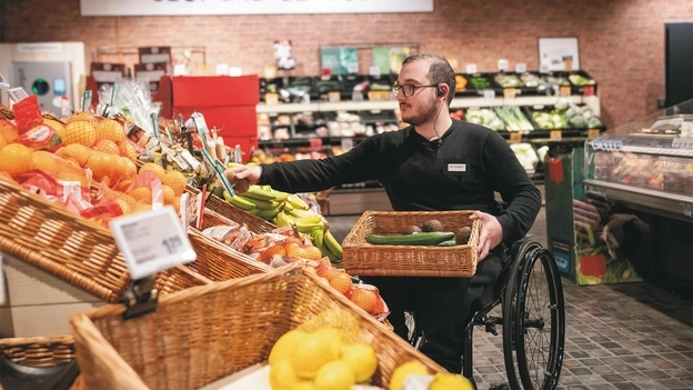 In einem Supermarkt-Gang prüft ein Mitarbeiter in schwarzer Hose und Pulli, der im Rolli sitzt,  Obst an einem Obststand. Aussortierte Ware legt er in einem Korb auf seinem Schoß.
